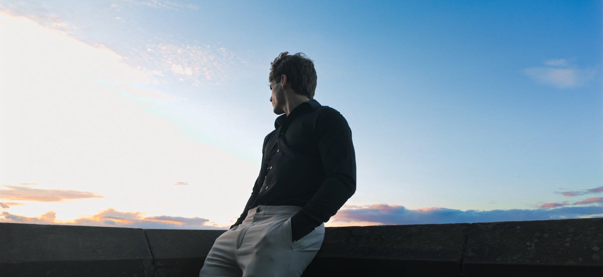 A man wearing a black long-sleeve button-up shirt stands outdoors against a scenic sunset sky, adjusting his sleeve.