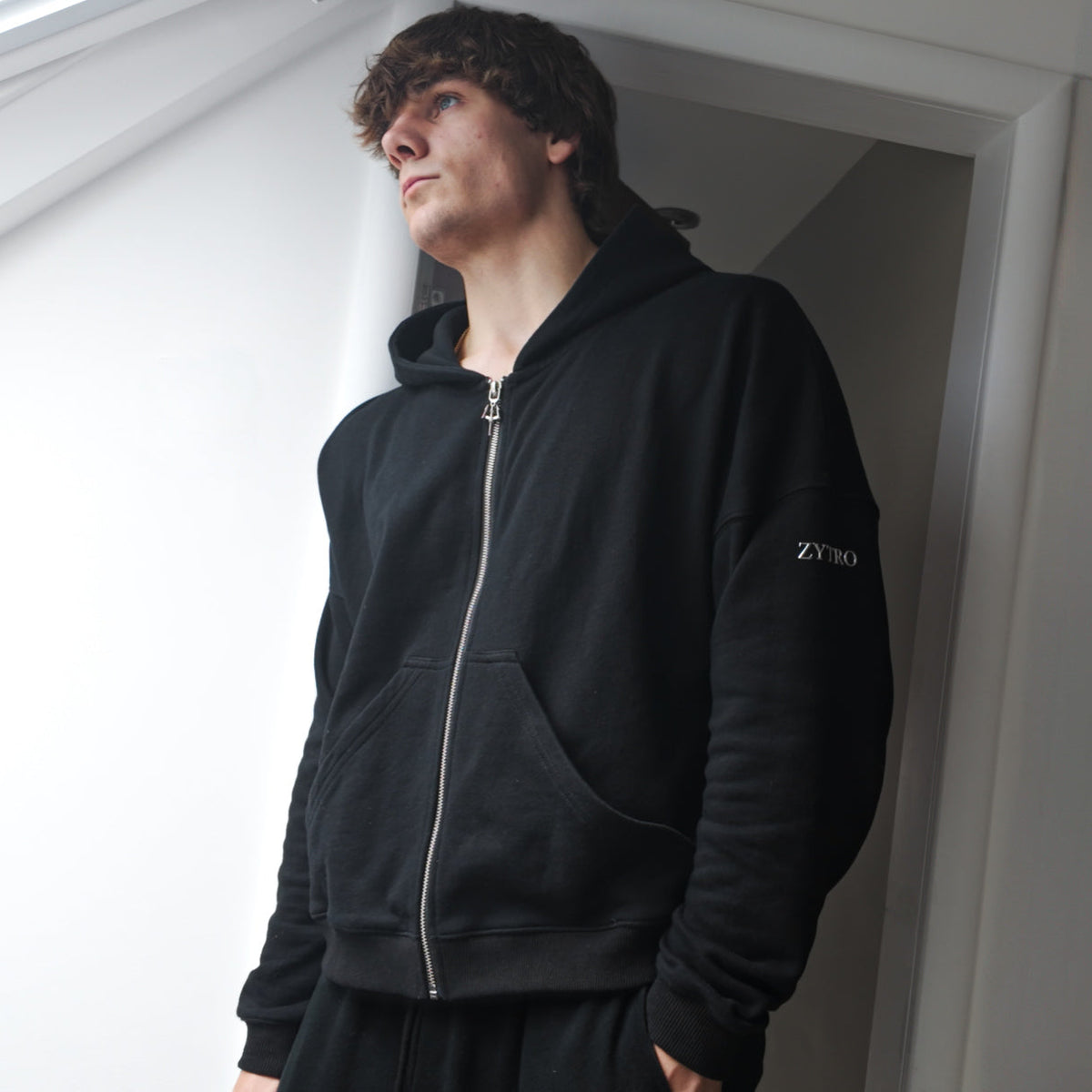 A man wearing a black zip-up hoodie with a silver zipper and a small silver "ZYTRO" logo on the left sleeve, standing in a doorway.