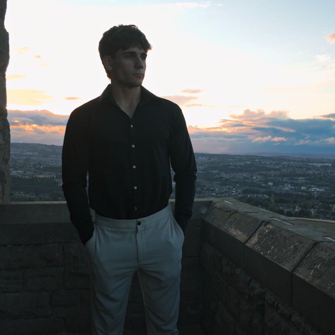 A man in a black shirt stands in profile looking out over a wide valley landscape at sunset, with hills, roads, and distant buildings visible below.